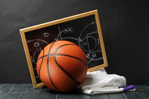 A basketball rests against a blackboard with tactical diagrams chalked on it, symbolizing a coachs strategic planning. The blackboard is in a wooden frame, and a white towel and colored chalks lie on the dark floor beside the ball.