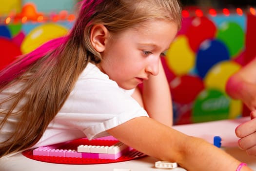 A young girl with curly hair, wearing a striped sleeveless dress, is focused on assembling a structure with vibrant toy blocks. The bright colors of the toys contrast with the blurred background, indicating an indoor play area.