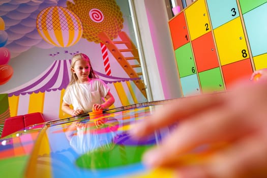 A joyful young girl is captured engaging in a playful board game, surrounded by bright and whimsical decorations in a colorful playroom setting. A hand is seen reaching for a game piece on the reflective, rainbow-hued surface, emphasizing the dynamic and interactive aspects of the game.