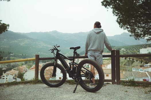 View from the back to a young active man cyclist standing near his electric battery powered bike, admiring beautiful mountains, enjoying riding in the nature