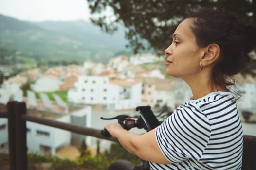 Rear view multi ethnic female cyclist relaxing on top of mountain, keeping hands on handlebar of his electric pedal-assist bicycle, enjoying amazing urban landscape below. Travel and active lifestyle