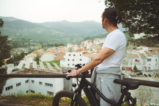 Rear view of a male cyclist in white t-shirt and gray sports shorts, having a break, standing by his electric bike, admiring the medieval village from the top of valley. Active healthy lifestyle.
