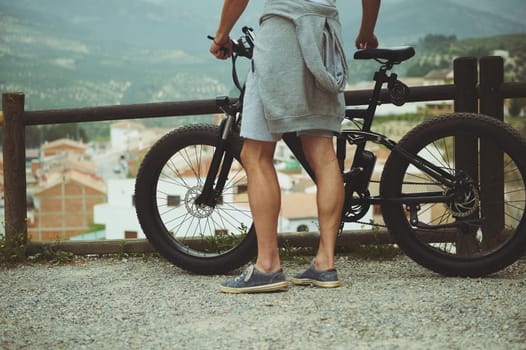 Close-up view from the back to a young active man cyclist standing near his electric battery powered bike, admiring beautiful mountains, enjoying riding in the nature. People. Freedom. Eco transport