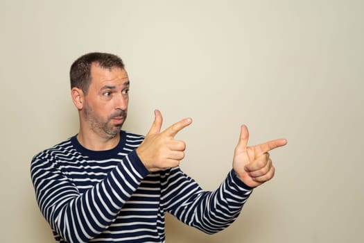Hispanic man with beard with two hands arms sign gesture pointing aside, isolated beige wall background. Feelings, signs and facial expression symbols of positive human emotions.