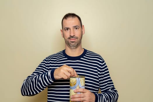A Hispanic man with a beard wearing a striped sweater putting a euro coin into a metal piggy bank, trying to save so he can travel on vacation. Isolated on beige studio background