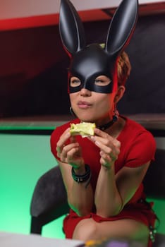 beautiful girl in bdsm rabbit mask and a bright red dress eats an apple in the kitchen in neon light promoting a healthy lifestyle vegetarianism
