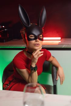 beautiful girl in a bdsm-style rabbit mask and a bright red dress with leather straps is posing sweetly in the kitchen in neon light