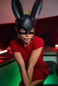 beautiful girl in a bdsm-style rabbit mask and a bright red dress with leather straps is posing sweetly in the kitchen in neon light