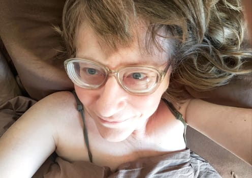 Shaggy cheerful middle aged woman with glasses smiling and resting in bed in early morning. Smiling Woman Relaxing on Brown Pillow