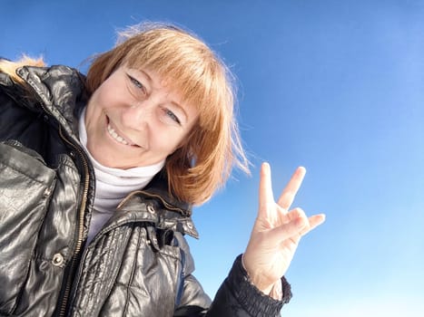 A cheerful middle aged woman in a winter coat taking selfie on nature outdoors in sunny day with blue sky