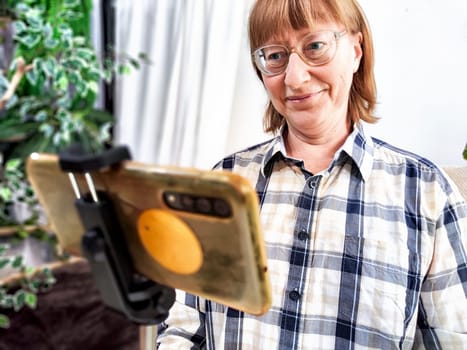 Smiling woman in glasses using a smartphone on a tripod for a video chat. A middle-aged woman posing and taking a selfie