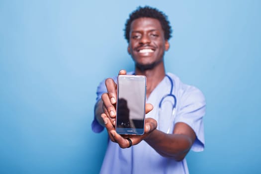 Male nurse looking at camera while carrying cell phone with blank screen, against blue background. Demonstrating medical expertise, black man wears scrubs and stethoscope while holding mobile device.