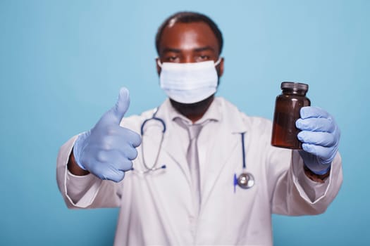 Portrait of medical specialist in a face mask, showing thumbs up while gripping pills bottle. Black doctor with stethoscope and gloves approving prescription painkillers with hand gestures.