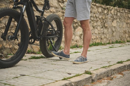 Close-up legs of a cyclist in sports shoes, pushing his bike while walking along the street. Man using e-bike as ecological sustainable mode of urban economic transport. People. Lifestyle. City life