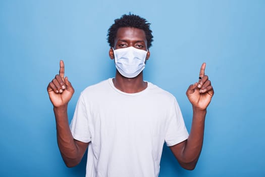 Portrait of black man wearing mask for coronavirus protection is pointing up with his two index fingers. African american guy wearing face mask while doing sign with hands in the studio.