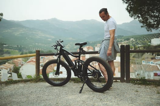 Caucasian young man standing with mountain bike on the top of hill, relaxing while cycling in the nature outdoors
