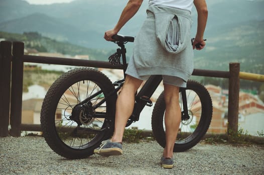 Cropped portrait of a young man cyclist, standing near his electric bike, having a break after biking in mountains. People. Active healthy lifestyle. E-bike as sustainable, eco friendly transportation