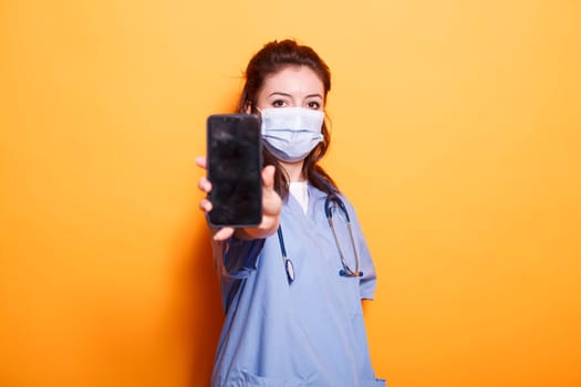 Nurse wearing scrubs and face mask, gripping cell phone with blank screen. Medical assistant stands in front of isolated background, looking at camera and displaying empty mobile device screen.