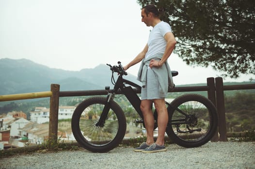Full length European male cyclist relaxing on top of mountain, keeping hands on handlebar of his electric pedal-assist bicycle, enjoying amazing urban landscape below. Travel and active lifestyle