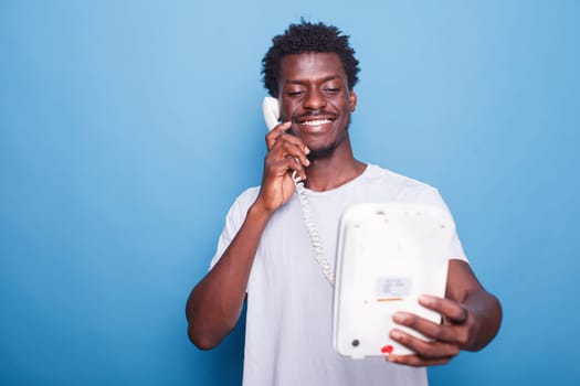 Young black man standing against isolated blue background is answering telephone call. Portrait of smiling african american guy enjoying a landline phone conversation with loved ones.