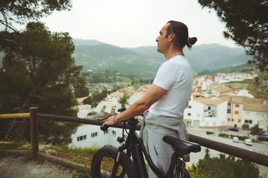 Side view of a male cyclist in white t-shirt and gray sports shorts, having a break, standing by his electric bike, admiring the medieval village from the top of valley