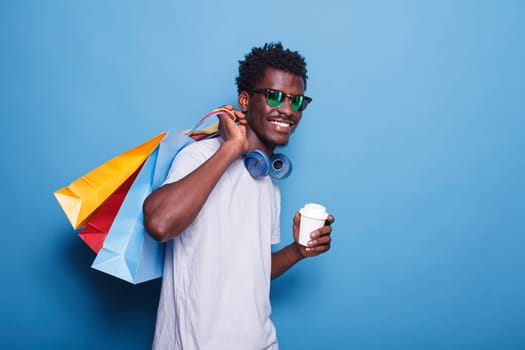 Joyful African American man holding a cup in happiness after a shopping spree. An upbeat male individual wearing sunglasses and headphones, clutching bags with products from Black Friday.