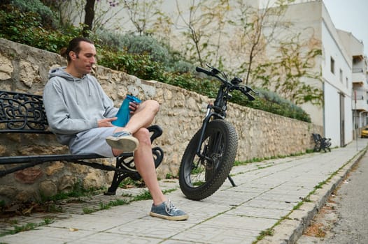Young adult sportsman, athletic man cyclist relaxing when riding electric bike in the city, holding a bottle of water while sitting on the bench. Urban lifestyle. Eco-friendly transport. Active people
