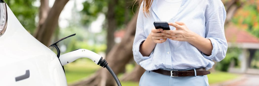 Panorama young woman recharge EV electric vehicle battery from EV charging station and using smartphone online banking to pay for electricity in city park. Eco friendly vehicle with EV car. Exalt