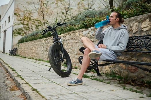 Full length portrait of Caucasian cyclist man drinking water, sitting on city bench. Electric motorbike, e-bike, electric bicycle bike on the foreground. Bike sharing city service. Sustainability.