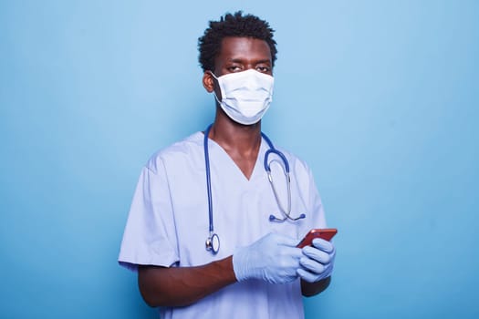 Close-up of nurse wearing face mask, holding smartphone for healthcare information during pandemic. African American medical professional stands confidently against blue background with mobile device.