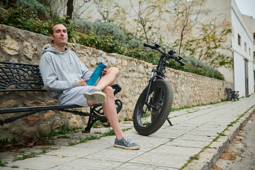 Young Caucasian man in gray hoodie and sports shorts, sitting on a bench near his rented electric bike, relaxing in the urban environment. Using e-bike as sustainable ecological mode of transport