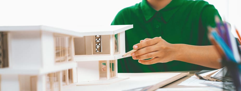 A cropped imaged of female designer measures a house model with a pencil, focusing intently on her work with house model and color swatches placed on table. Focus on hand. Closeup. Variegated.