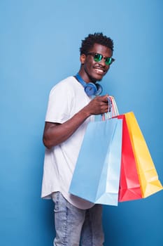 African American guy wearing sunglasses, holding shopping bags, and standing in front of an isolated blue background. Black man with wireless headset on his neck, carrying colorful parcels.