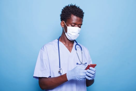 African american medical assistant in blue scrubs grasping smartphone in hand. Male healthcare specialist wearing face mask and gloves for covid 19 protection while having a mobile device.