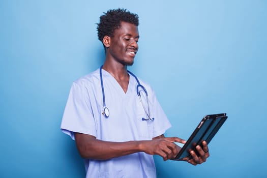 Smiling black doctor with stethoscope is using a tablet for healthcare research. African American medical professional surfing the net on his digital device, standing against isolated background.