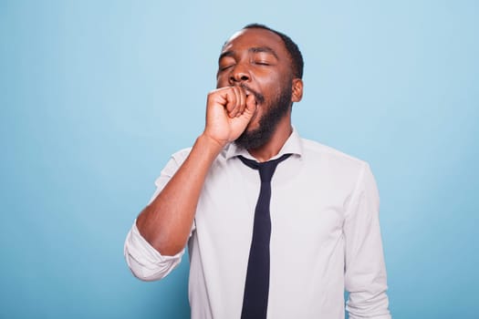 Tired black man yawning with his mouth covered in front of isolated background. Exhausted African American male entrepreneur looking at camera, feeling fatigued and overworked.