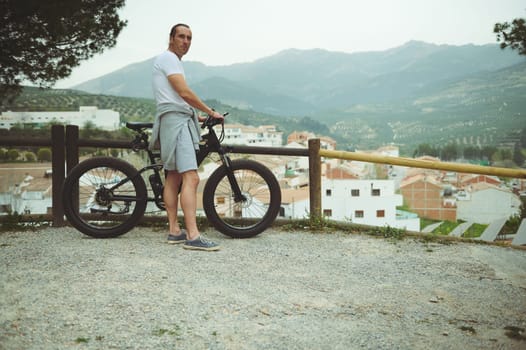 European man cyclist admiring amazing nature standing on top of hill with his motor-powered booster mountain bike. People. Active healthy lifestyle. Sport. Achievements. Inspired by nature. Copy space