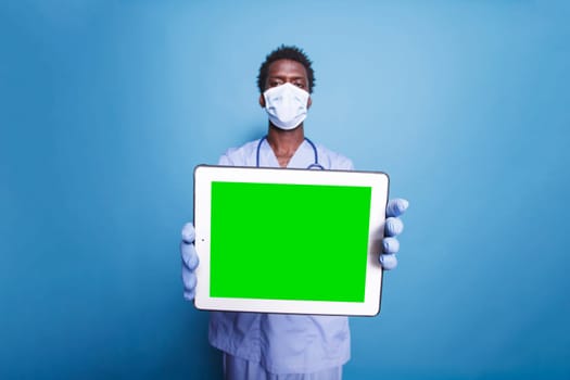 Male healthcare professional stands with a tablet displaying blank green screen while wearing scrubs and face mask. Holding digital device with chromakey template is a black man.