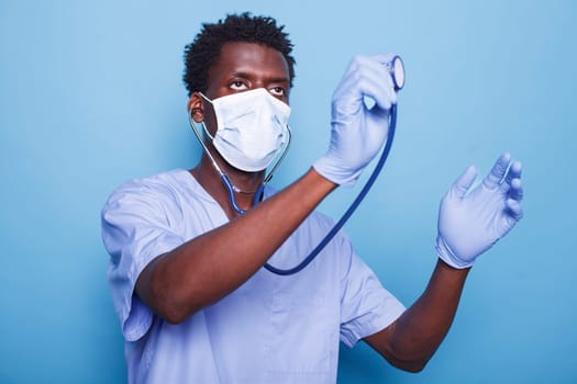 Male healthcare nurse is holding stethoscope to examine cardiology patient heartbeat. African American man wearing gloves and face mask as cardiologist fights coronavirus epidemic.