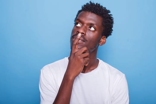 Portrait of black man brainstorming and pondering while grasping his chin. In studio, focused African American guy with hand on face, thinking and wondering in front of blue background.