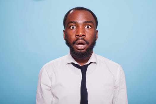 Stunned businessman stands in front of an unbelievable situation on a blue background, his mouth hanging open in shock. Surprised black individual with wide eyes and a jaw drop.