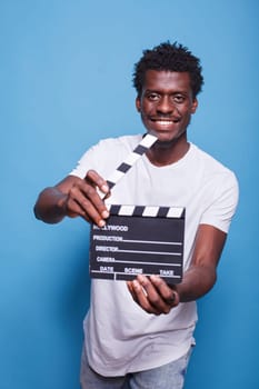 African american filmmaker holding open clapper in hands looking confident with a smile on his face. Black movie producer with clapperboard standing proud in front of isolated blue background.