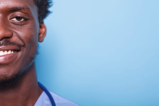 Close-up portrait of cheerful male nurse wearing scrubs and stethoscope on a blue background. Half face, cropped image of confident African American medical physician smiling at camera.