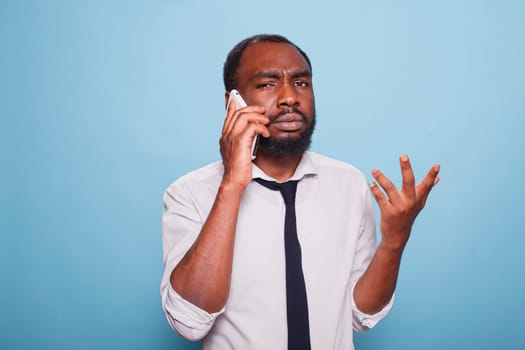 Frowning male freelancer explaining problem on smartphone doing hand gesture against blue background. Upset african american businessman in bad mood trying to understand business conversation.