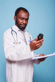 Dedicated male pharmacist grasping a clipboard while examining prescription pill bottle. Black man dressed in lab coat attentively inspecting medicine bottle while standing against blue background.