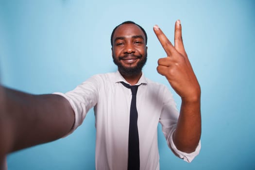 POV of young male entrepreneur making online vlog for social media advertising. Black businessman taking a selfie with peace sign hand gesture in front of isolated blue background.