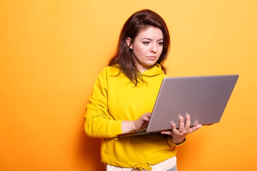 Female freelancer utilizing modern technology by using digital laptop. Portrait of focused woman working online on wireless computer while standing over isolated orange background.