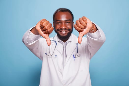 Black man wearing a hospital uniform and stethoscope standing and doing thumbs down to the camera. Portrait of african american medical specialist making disapproval hand gestures.