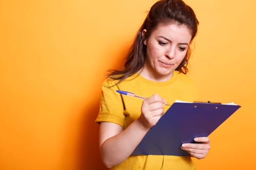 Portrait of female freelancer going through her documents and checking off items on clipboard. Caucasian lady standing in front of isolated background while using a pen to write notes.