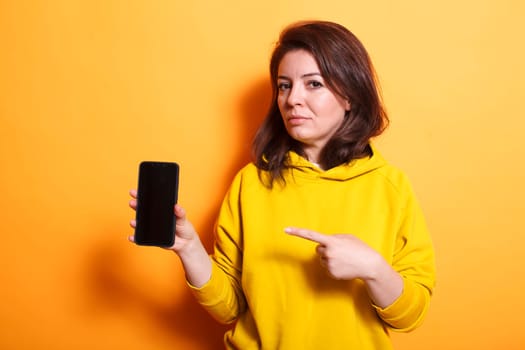 Brunette lady showing cellphone with empty screen at camera in studio. Woman pointing at mobile phone with isolated mockup template on display. Person standing over orange background